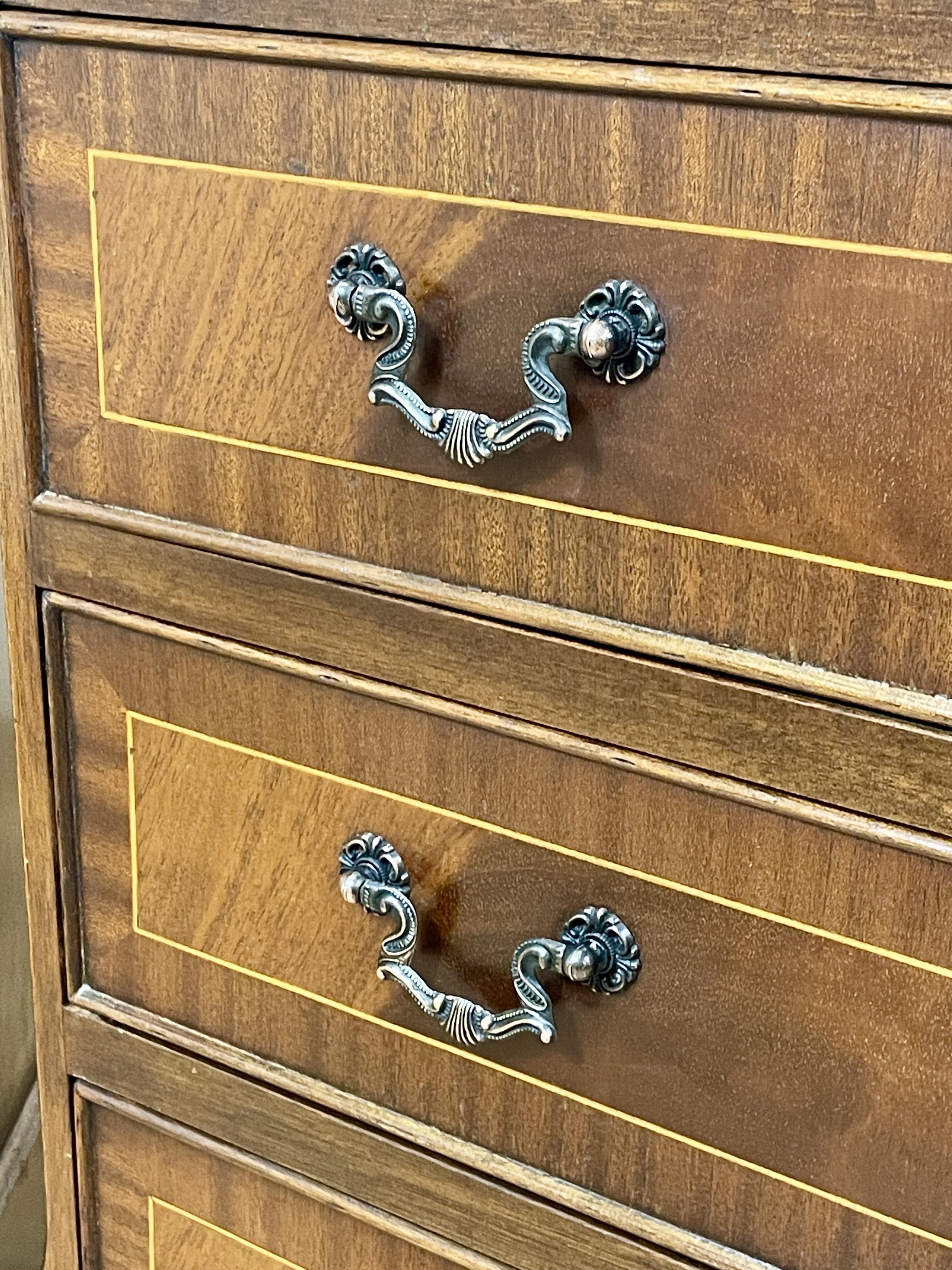 Vintage pair of mahogany bedside chest of drawers with five drawers each, featuring decorative inlaid tops and brass handles, displayed against a cream wall on a wooden floor.