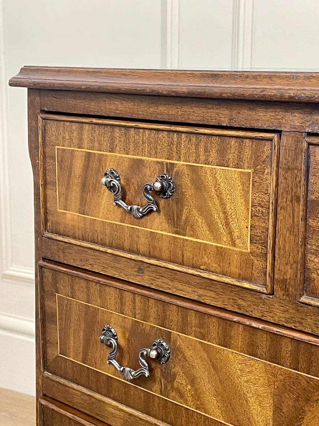 Vintage pair of mahogany bedside chest of drawers with five drawers each, featuring decorative inlaid tops and brass handles, displayed against a cream wall on a wooden floor.