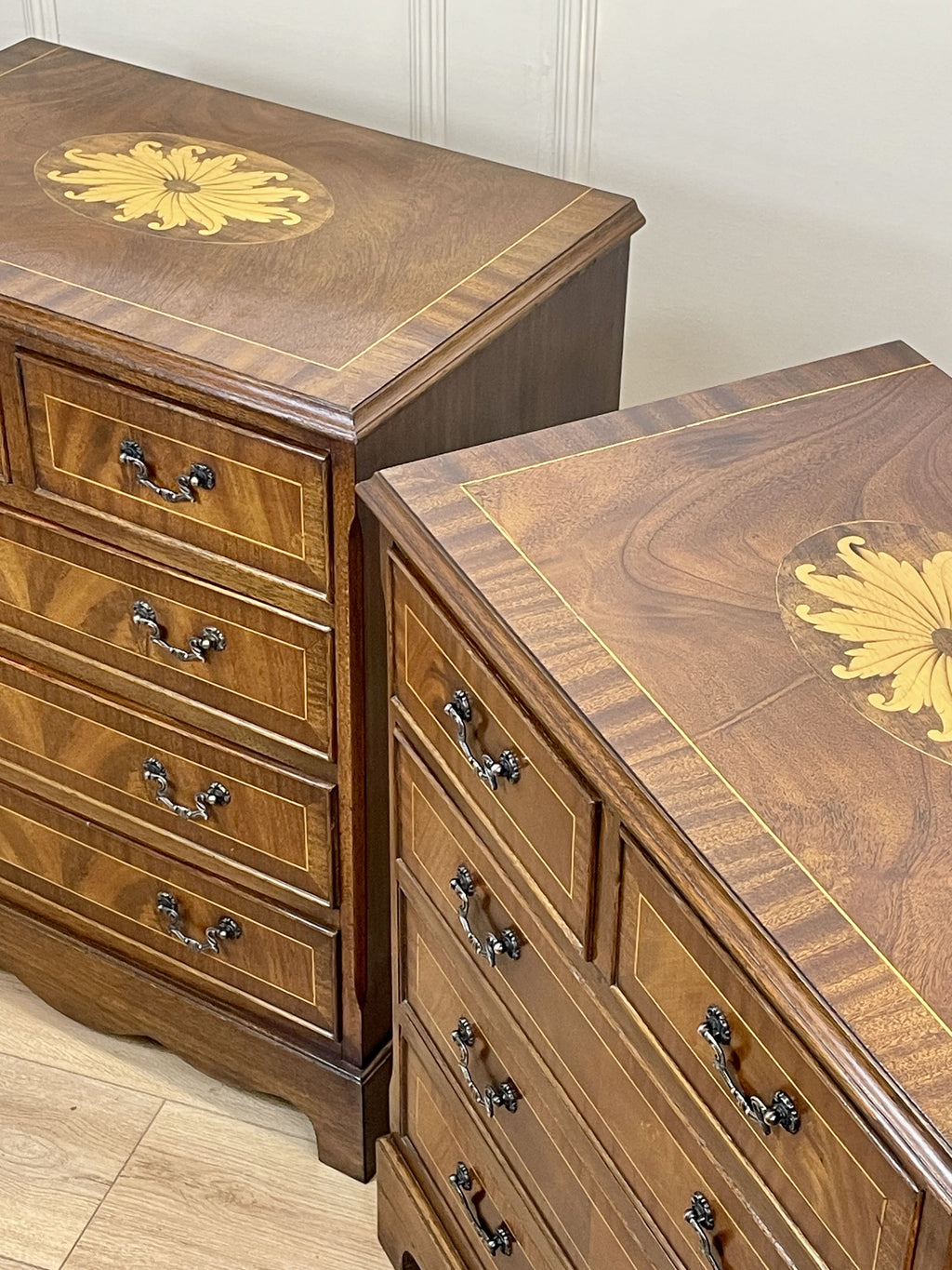 Vintage pair of mahogany bedside chest of drawers with five drawers each, featuring decorative inlaid tops and brass handles, displayed against a cream wall on a wooden floor.
