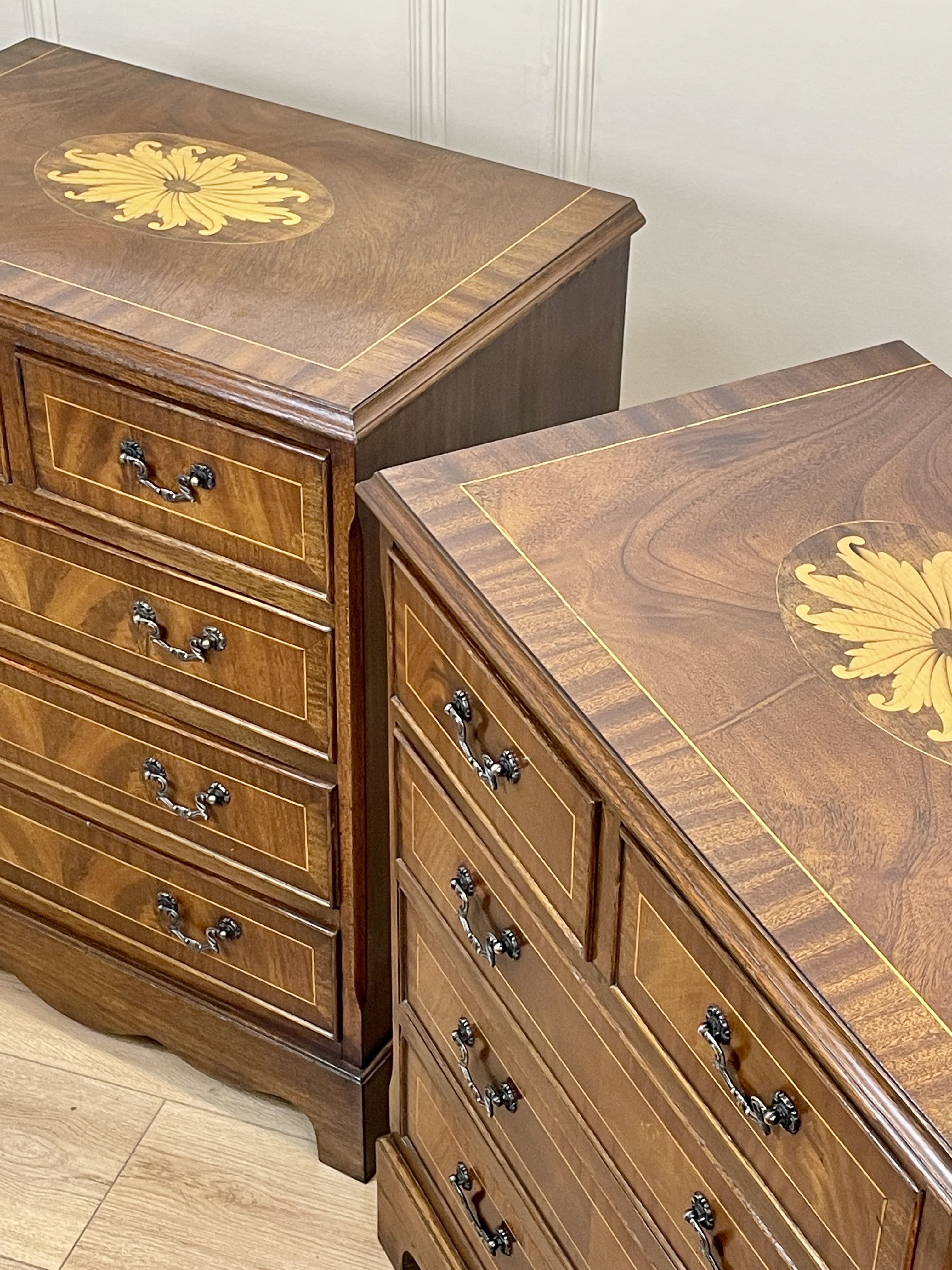 Vintage pair of mahogany bedside chest of drawers with five drawers each, featuring decorative inlaid tops and brass handles, displayed against a cream wall on a wooden floor.