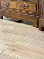 Vintage pair of mahogany bedside chest of drawers with five drawers each, featuring decorative inlaid tops and brass handles, displayed against a cream wall on a wooden floor.
