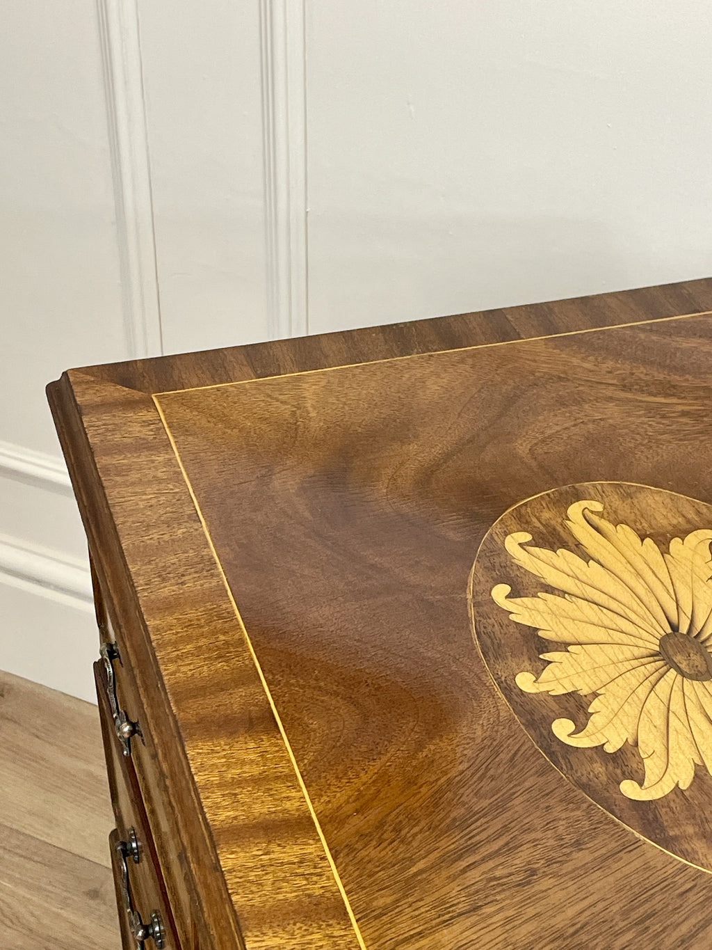 Vintage pair of mahogany bedside chest of drawers with five drawers each, featuring decorative inlaid tops and brass handles, displayed against a cream wall on a wooden floor.