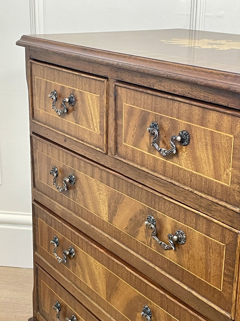 Vintage pair of mahogany bedside chest of drawers with five drawers each, featuring decorative inlaid tops and brass handles, displayed against a cream wall on a wooden floor.