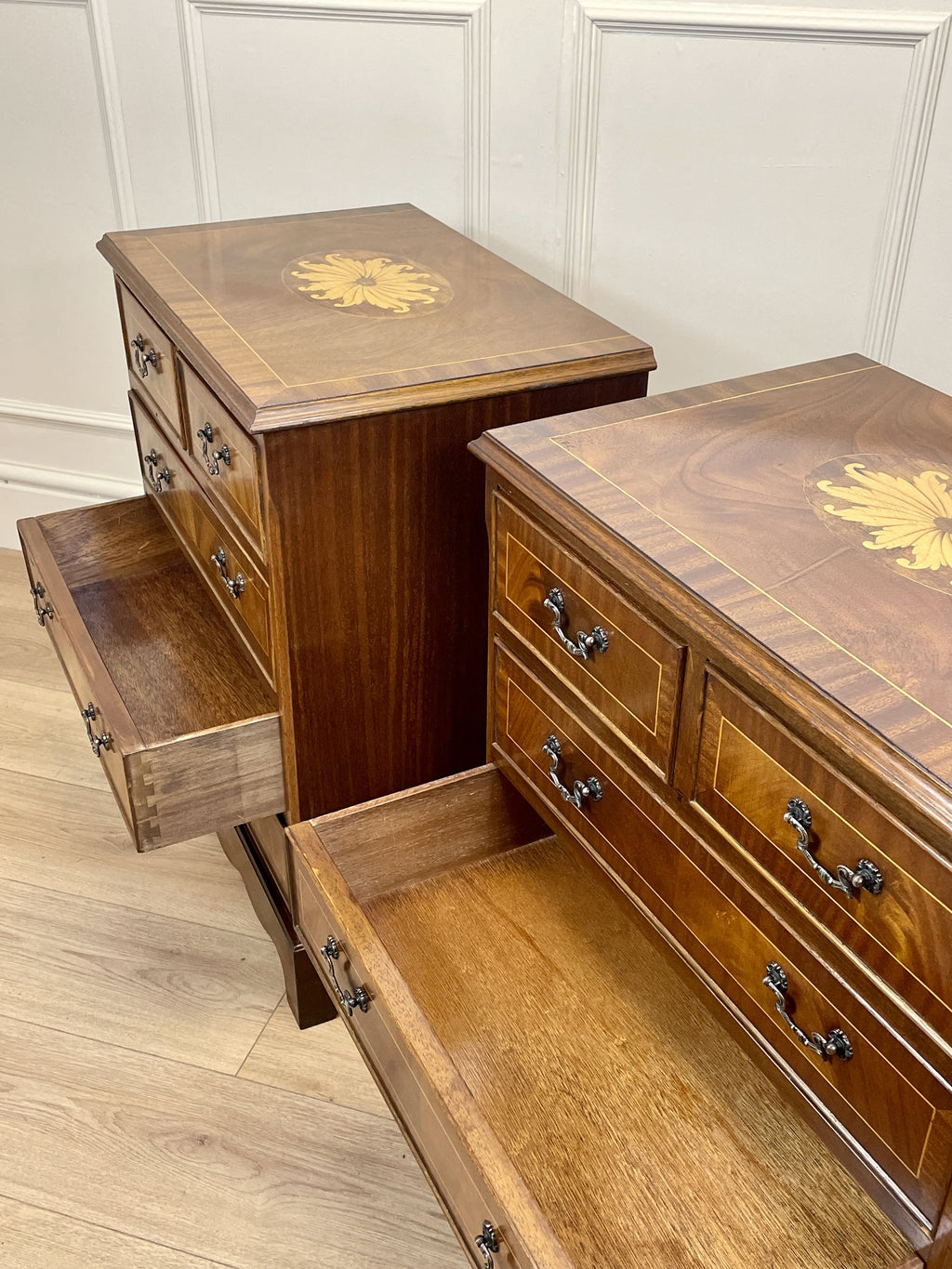Vintage pair of mahogany bedside chest of drawers with five drawers each, featuring decorative inlaid tops and brass handles, displayed against a cream wall on a wooden floor.