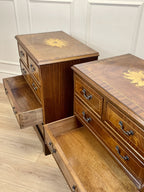 Vintage pair of mahogany bedside chest of drawers with five drawers each, featuring decorative inlaid tops and brass handles, displayed against a cream wall on a wooden floor.