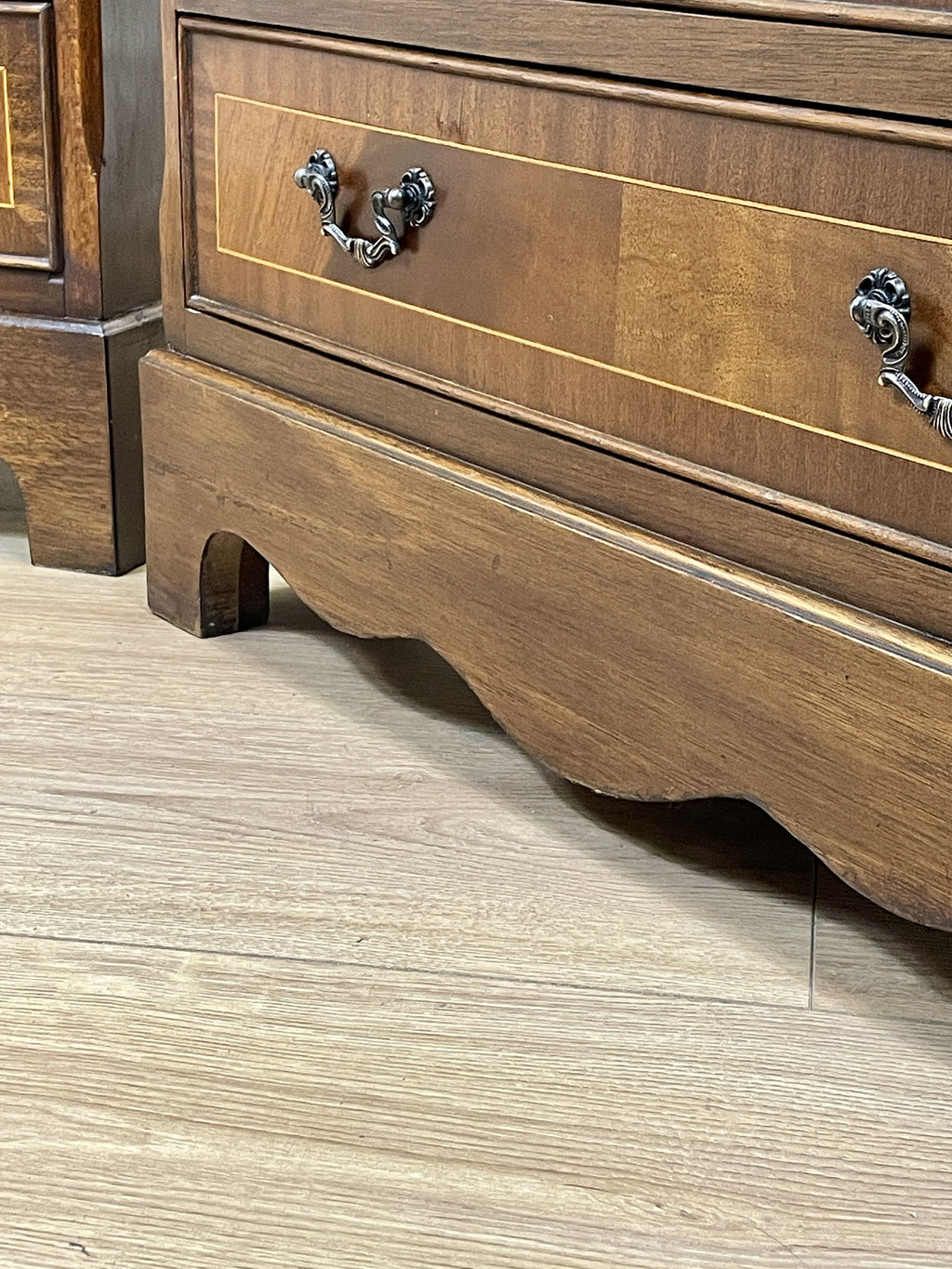Vintage pair of mahogany bedside chest of drawers with five drawers each, featuring decorative inlaid tops and brass handles, displayed against a cream wall on a wooden floor.