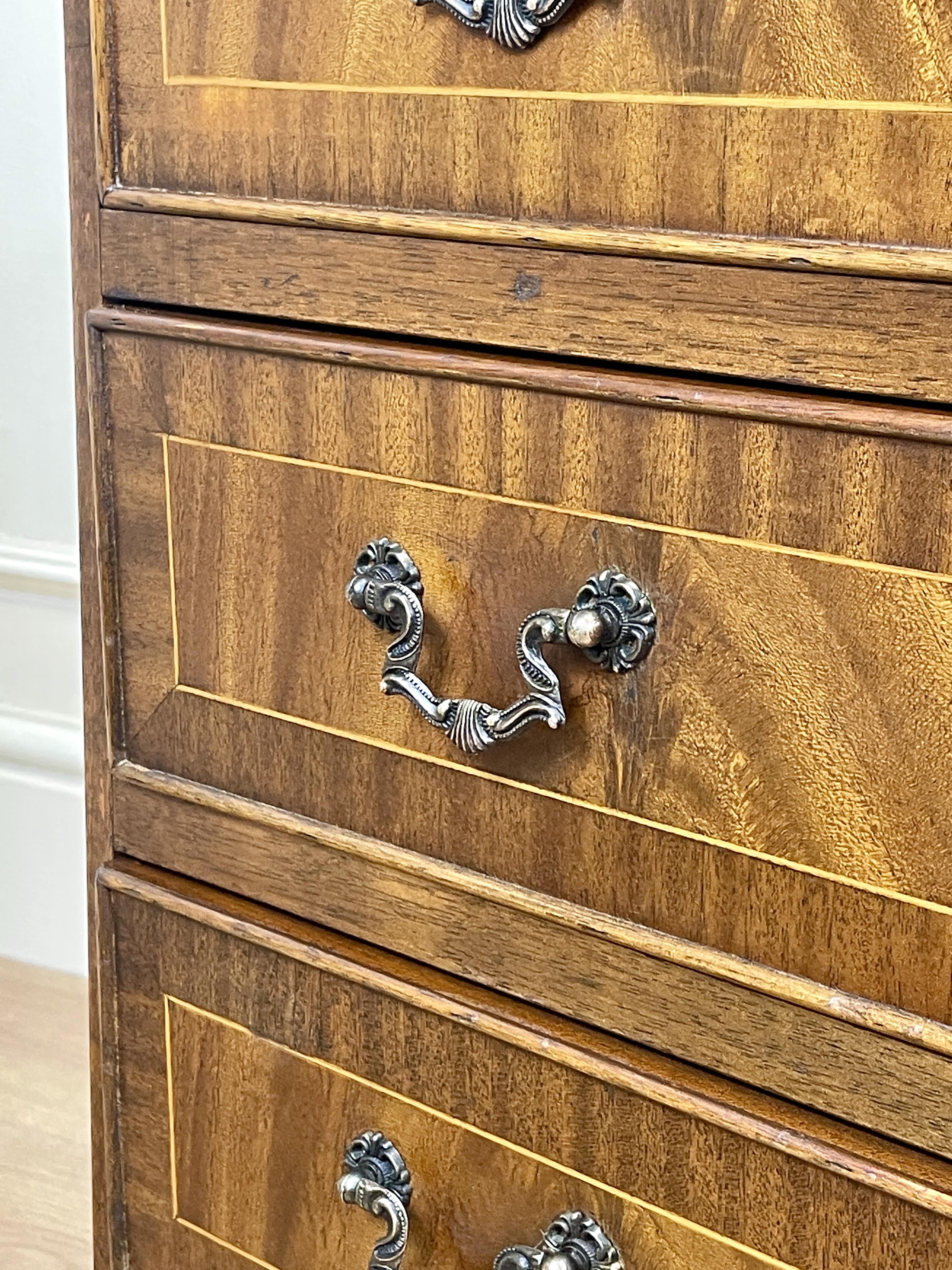 Vintage pair of mahogany bedside chest of drawers with five drawers each, featuring decorative inlaid tops and brass handles, displayed against a cream wall on a wooden floor.