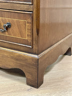 Vintage pair of mahogany bedside chest of drawers with five drawers each, featuring decorative inlaid tops and brass handles, displayed against a cream wall on a wooden floor.
