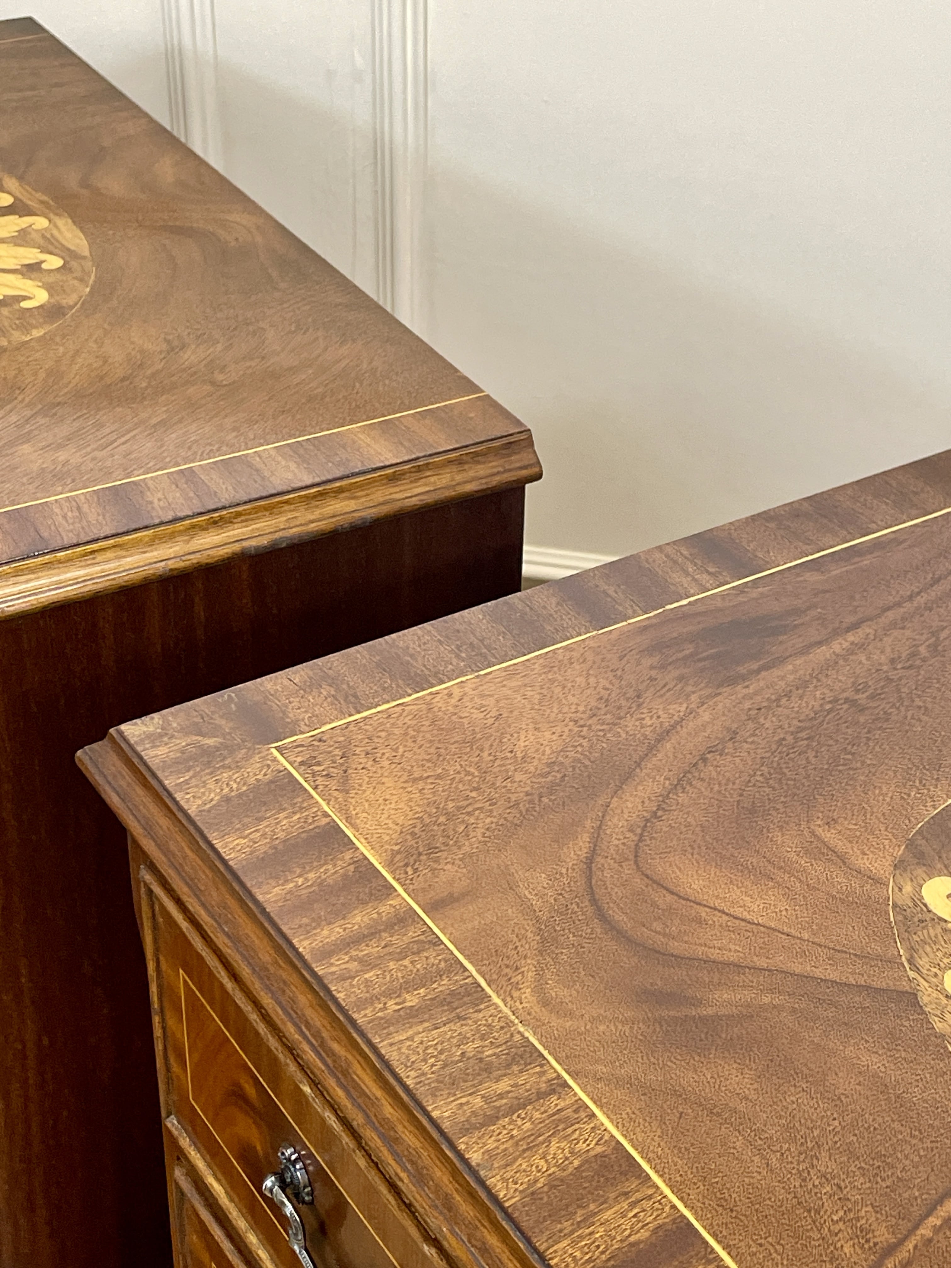 Vintage pair of mahogany bedside chest of drawers with five drawers each, featuring decorative inlaid tops and brass handles, displayed against a cream wall on a wooden floor.