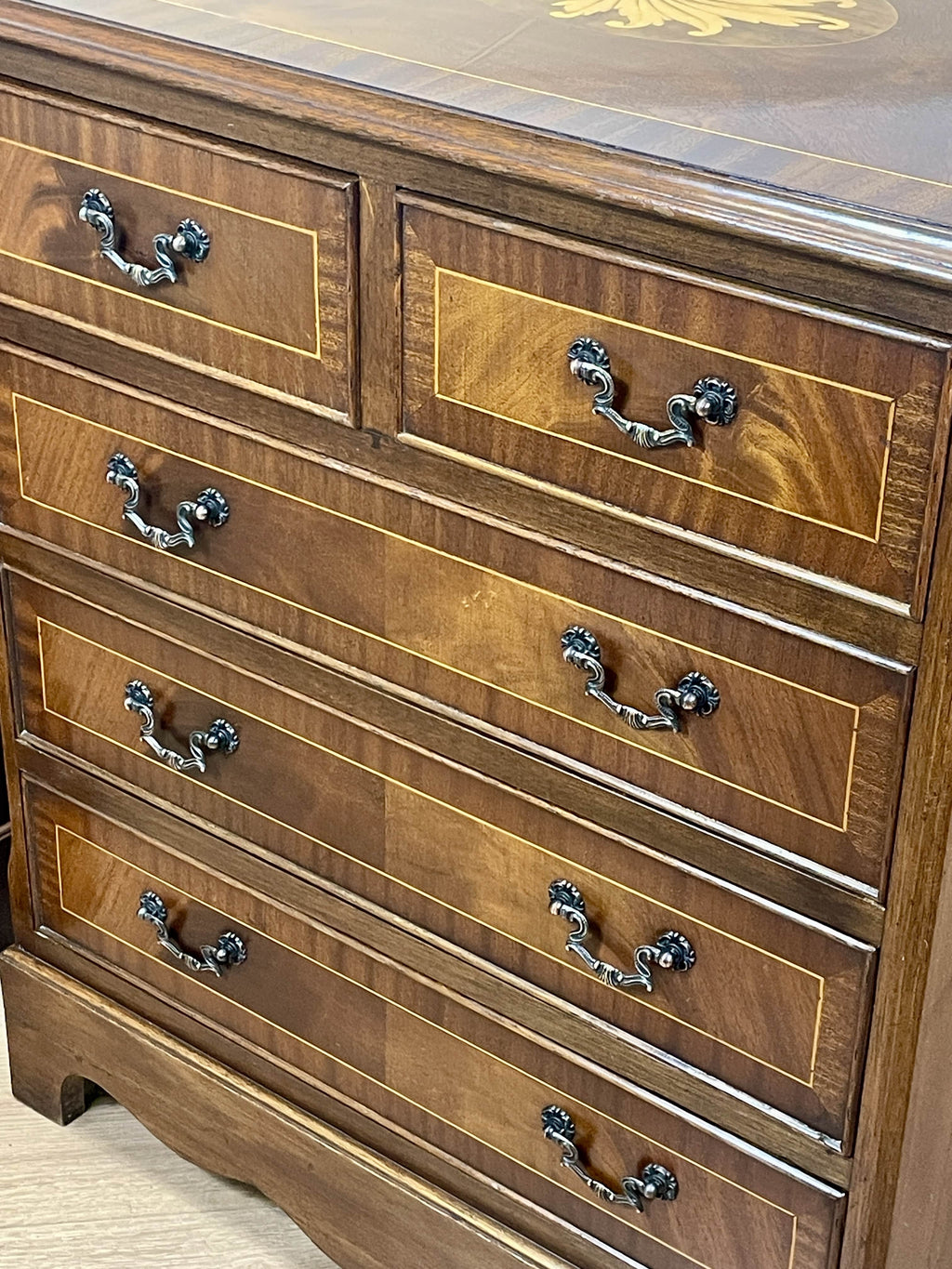Vintage pair of mahogany bedside chest of drawers with five drawers each, featuring decorative inlaid tops and brass handles, displayed against a cream wall on a wooden floor.