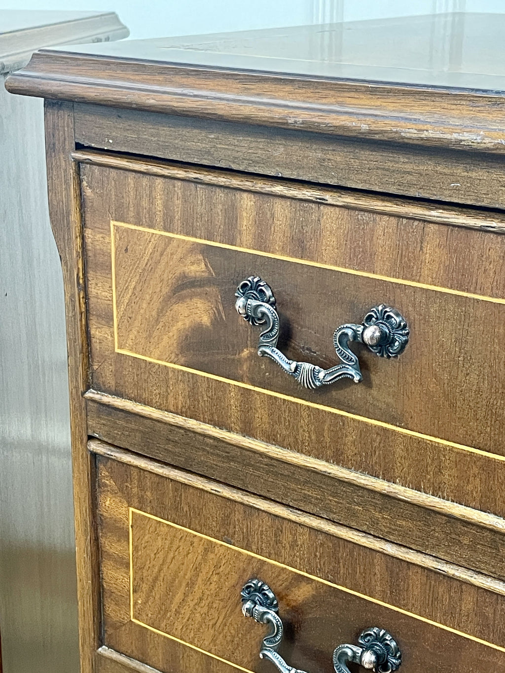 Vintage pair of mahogany bedside chest of drawers with five drawers each, featuring decorative inlaid tops and brass handles, displayed against a cream wall on a wooden floor.