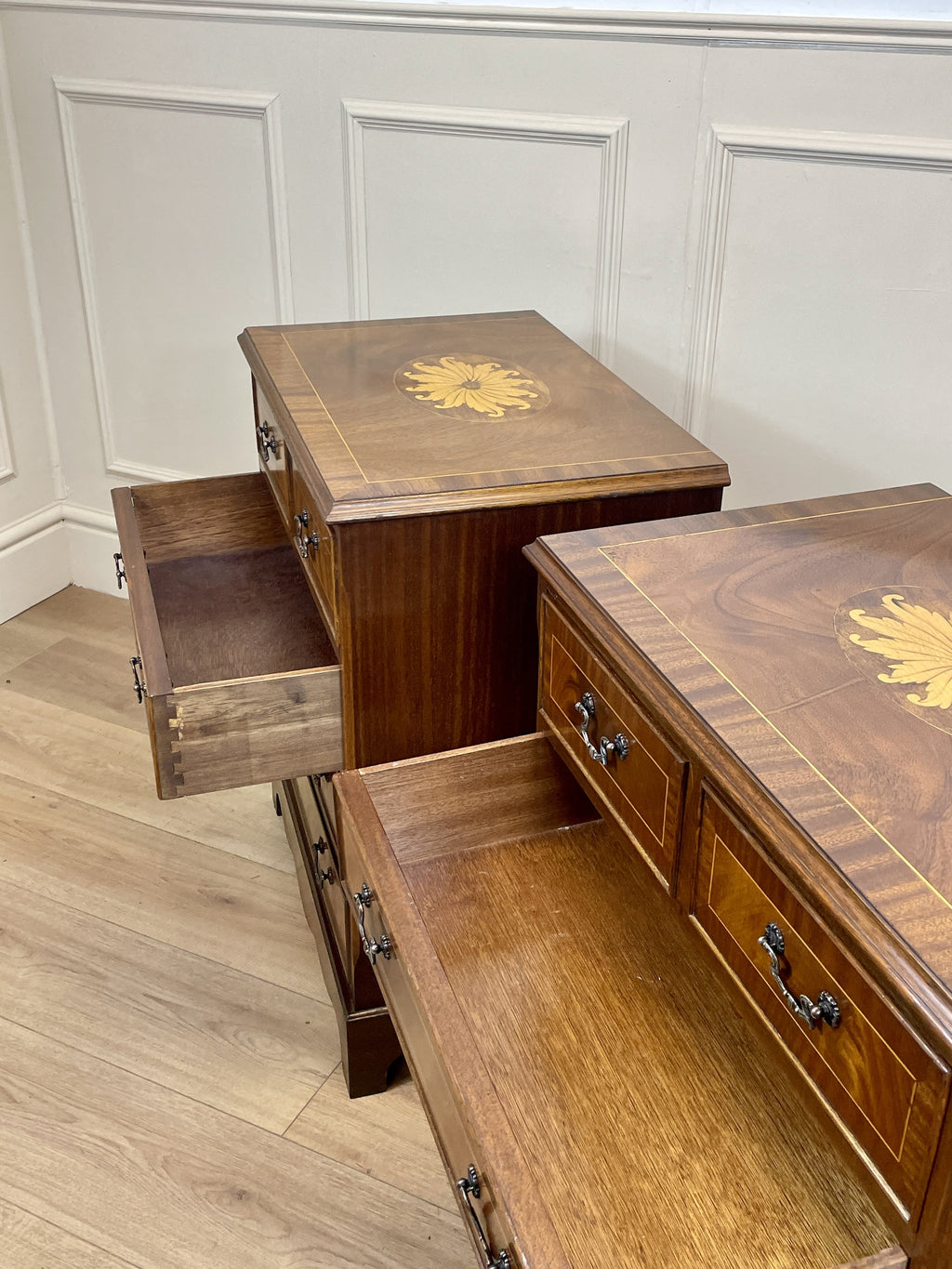 Vintage pair of mahogany bedside chest of drawers with five drawers each, featuring decorative inlaid tops and brass handles, displayed against a cream wall on a wooden floor.