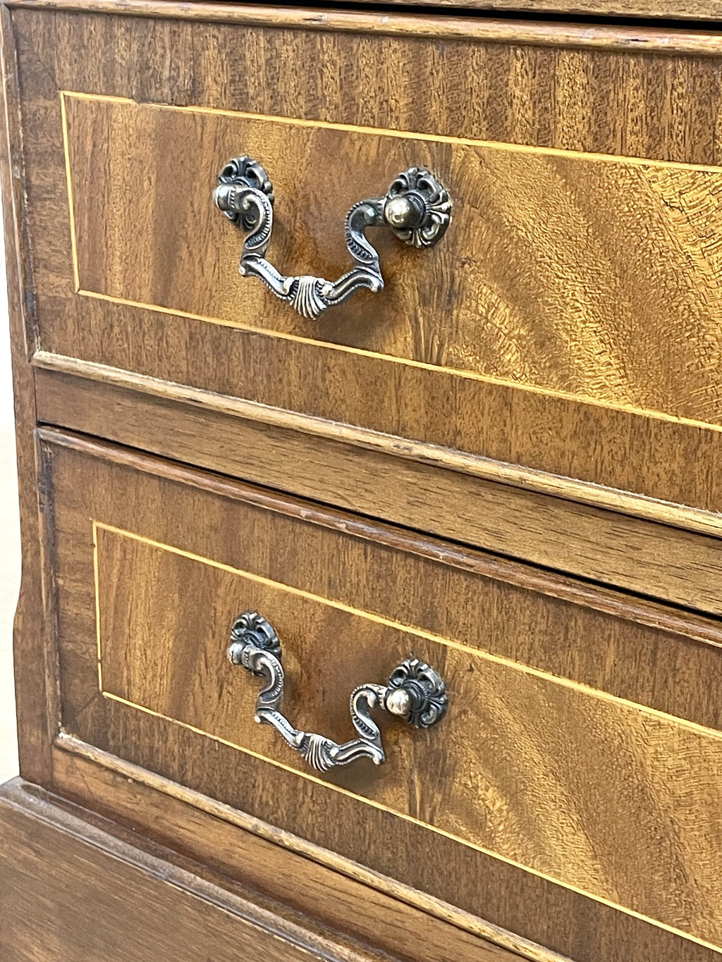 Vintage pair of mahogany bedside chest of drawers with five drawers each, featuring decorative inlaid tops and brass handles, displayed against a cream wall on a wooden floor.