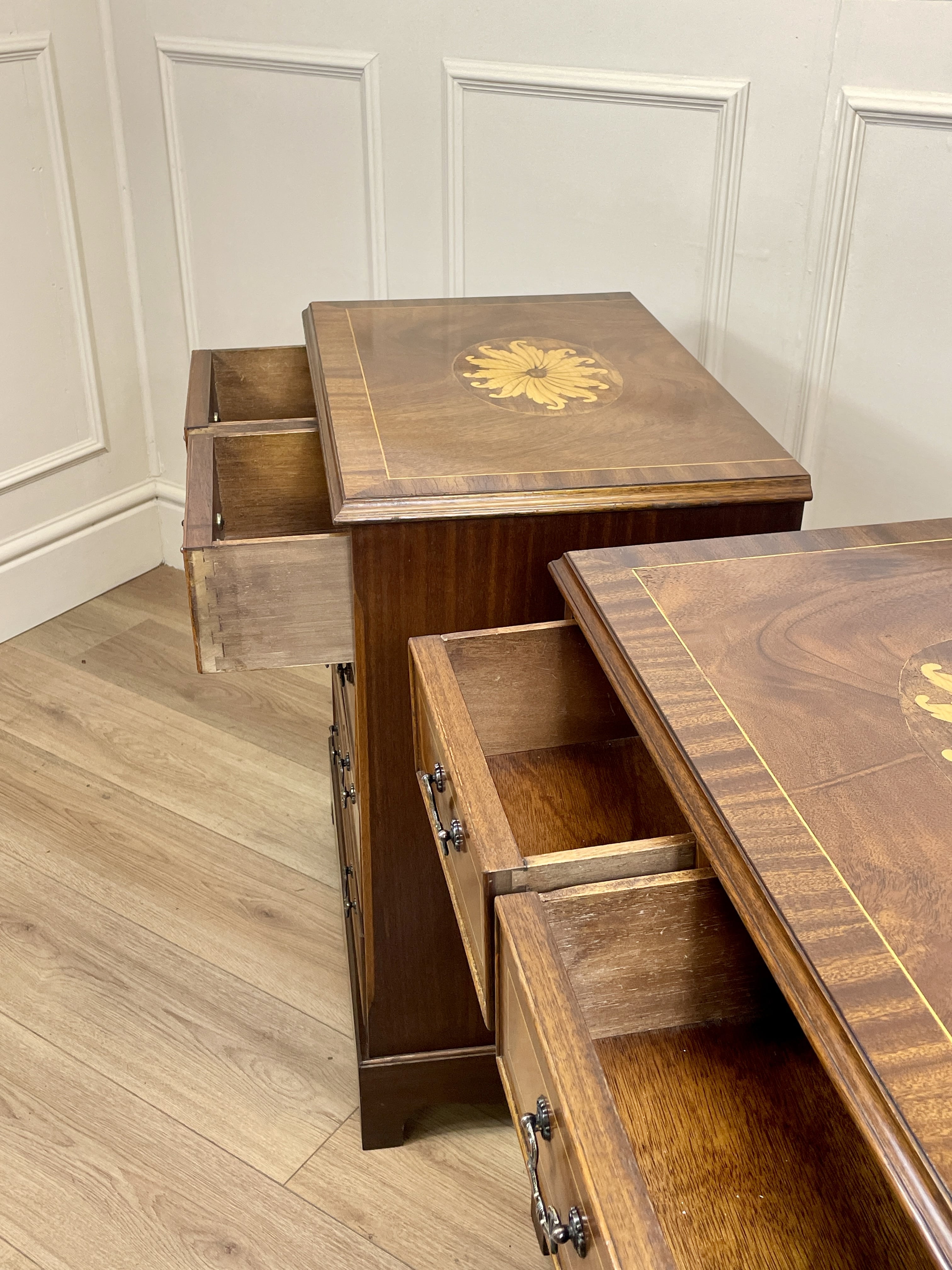 Vintage pair of mahogany bedside chest of drawers with five drawers each, featuring decorative inlaid tops and brass handles, displayed against a cream wall on a wooden floor.
