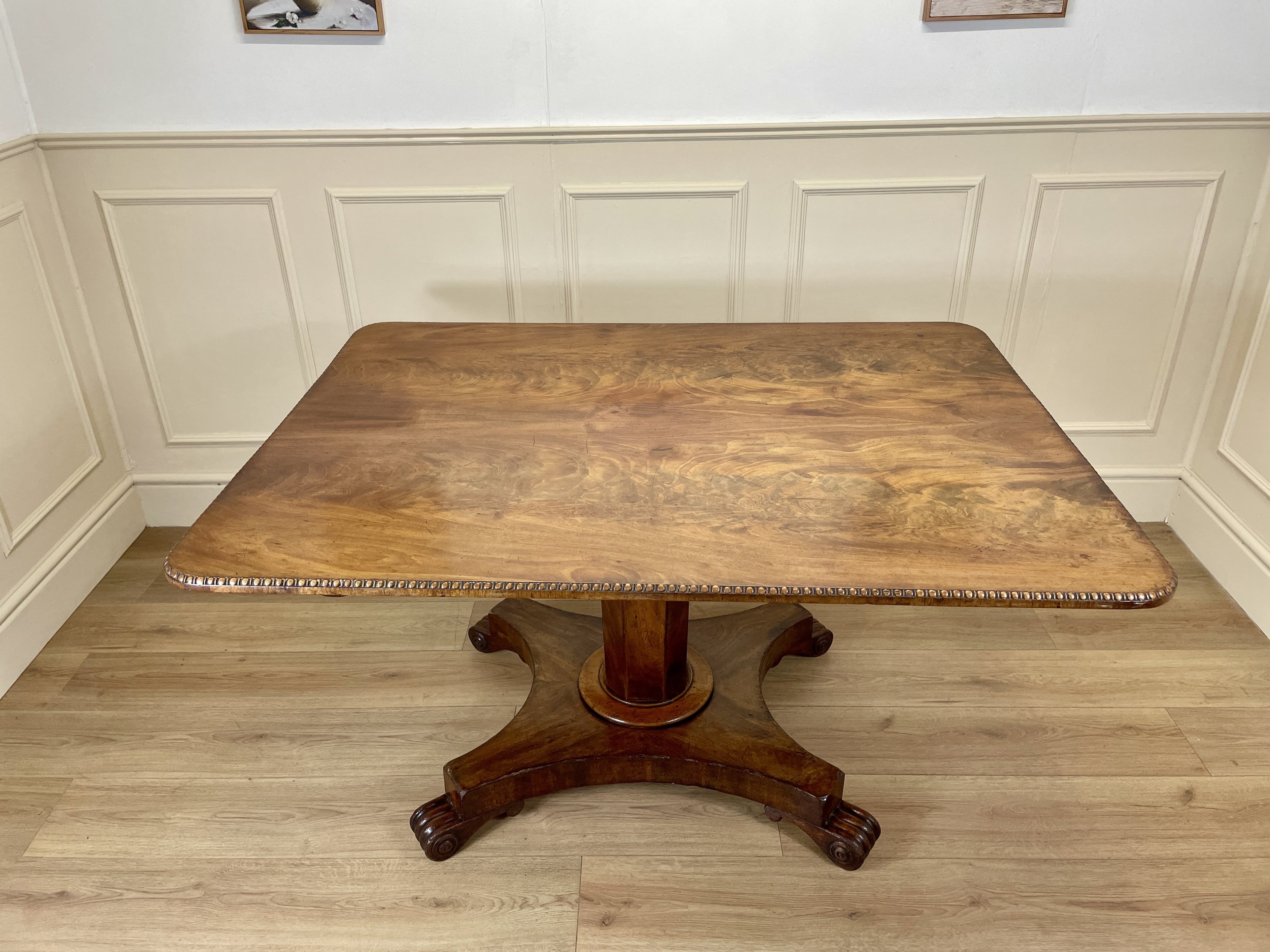 Wooden table with a round base in a room with white paneled walls and wooden floor.
